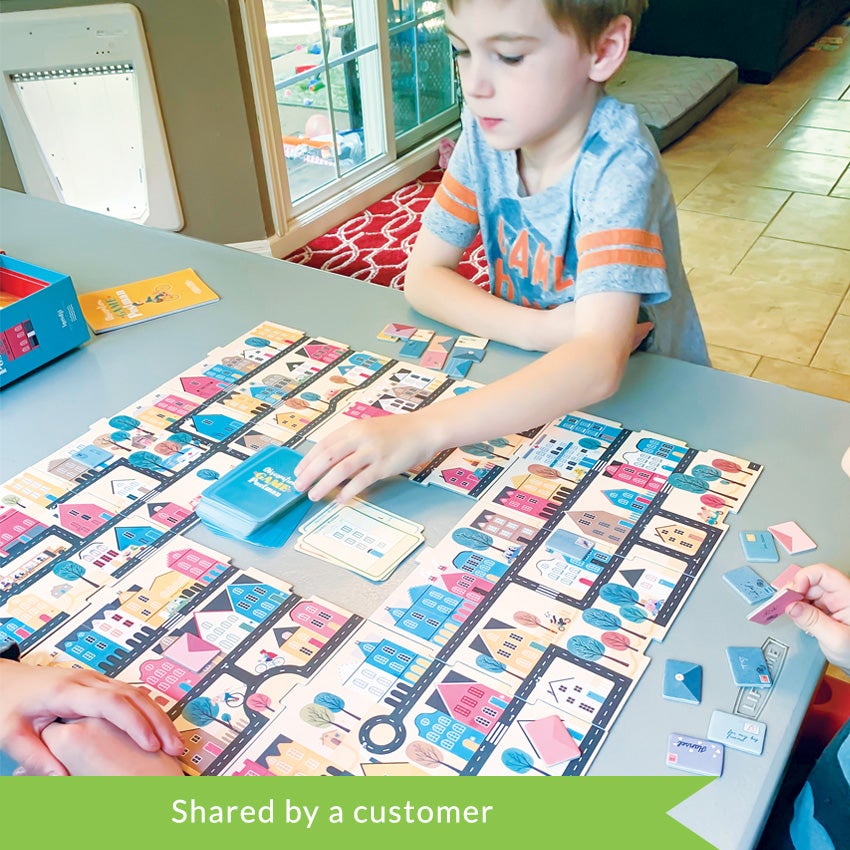 A customer photo of 3 children playing the Postman Observation game. The large board on the table has a square missing in the middle with 2 stacks of cards inside; 1 blue, 1 white. The board is covered in colorful town buildings and houses, roads and trees. On the sides of the board are some cut-out postal letter pieces. There is a boy at the top that is reaching out to grab a blue card. You can see only the hands of the other 2 children playing in the lower-left and lower-right.