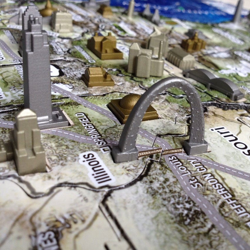A close up of a the U S A History Over Time Puzzle. The section focuses on Illinois and the Saint Louis Arch. You can see many other gold, silver, and bronze colored buildings. The puzzle base has different types of ground, waterways, and roads. In the middle is Springfield.