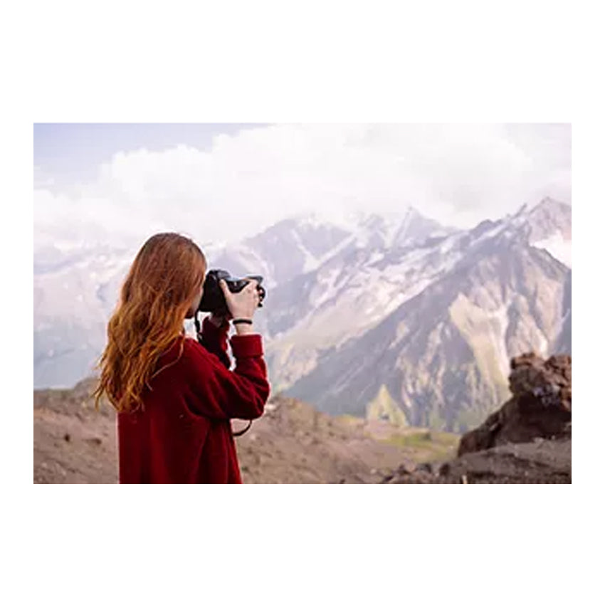 A photo of a red-haired teenager in a burgundy shirt holding up a large digital camera taking a picture of the mountainous scenery.