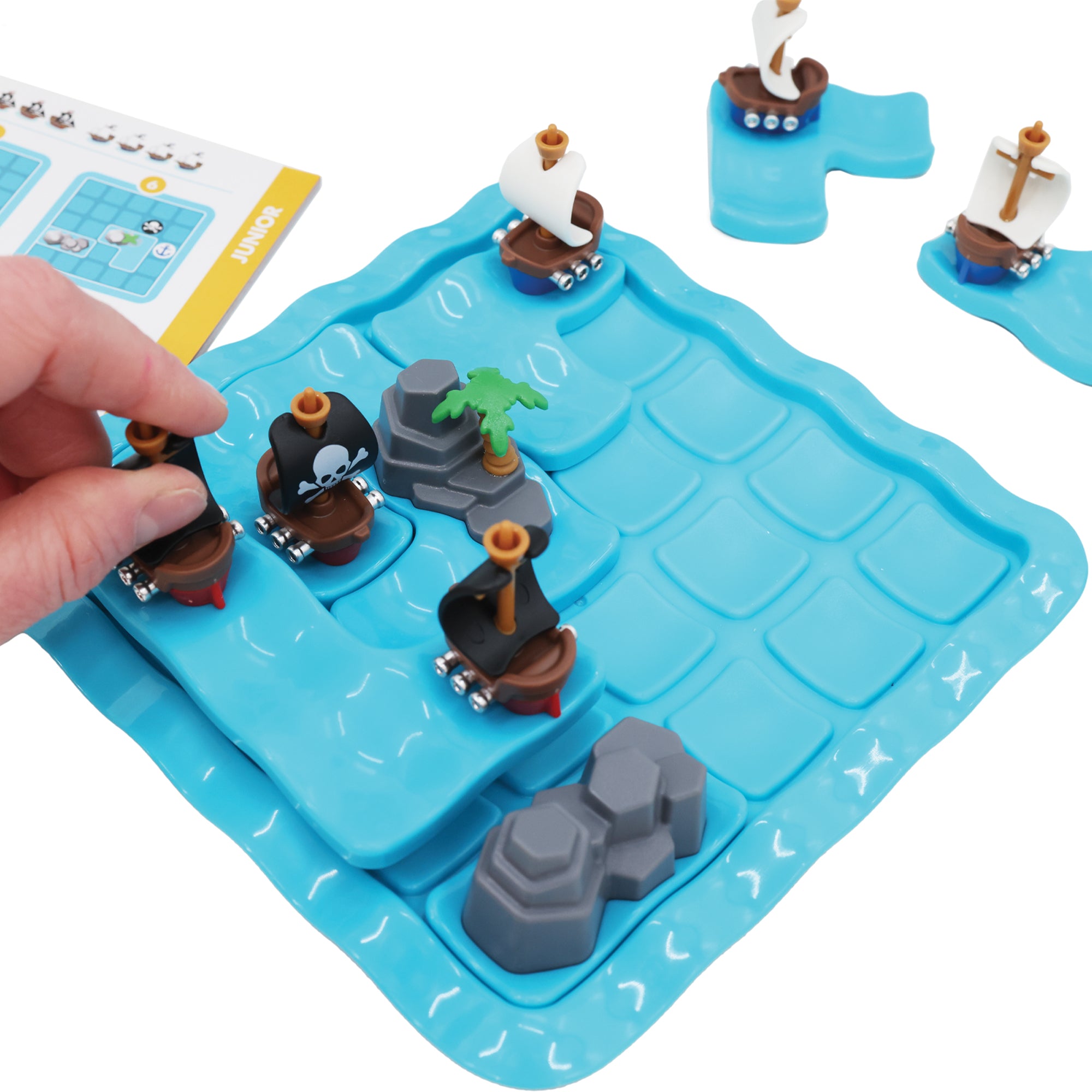 A hand putting a ship piece in place on the Pirates Crossfire game board. The gameboard is blue and wavy in a square shape with rounded corners. There are 4 ship and rock pieces set in place on the board. There are 2 ship pieces off the board to the top. On the left is the instruction booklet open to a junior puzzle.