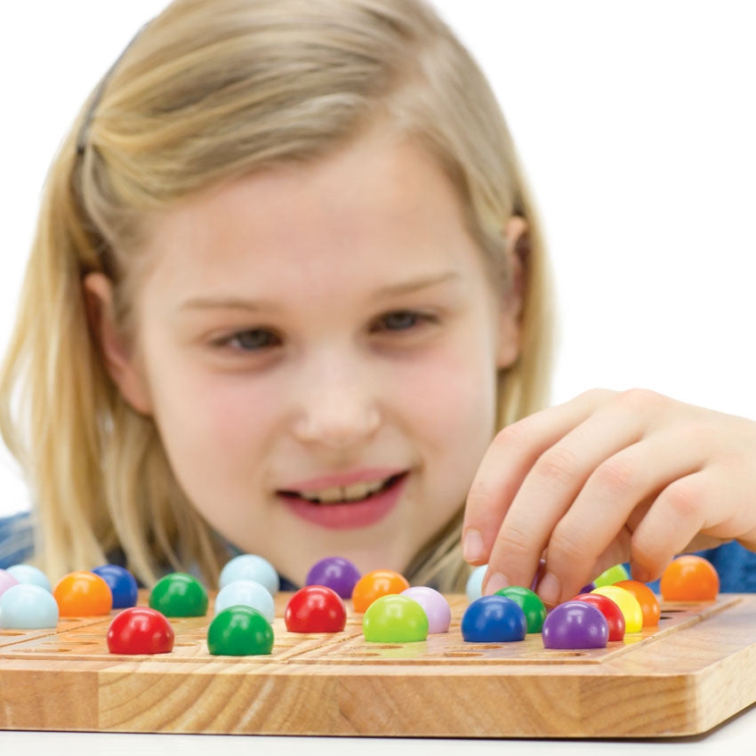 A close up of a blonde girl is in the middle of playing ColorKu. She is looking down and smiling at the board in front of her. The board in front of her has many pieces in place on top. She is placing a piece on the board with her left hand. The colored balls are red, orange, yellow, bright green, dark green, light blue, dark blue, light purple, and dark purple.