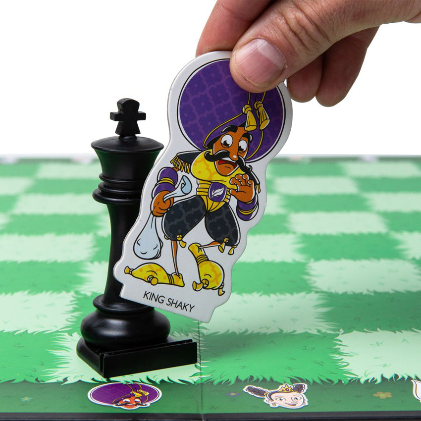 A close up of a hand coming out from the top-right corner and holding onto a Story Time Chess game piece. The cut-out piece is of “King Shaky.” He is wearing a balloon style outfit of purple, gold, and black. He has a white bag in his right hand and is looking down with a scared look on his face and his other hand protecting his face. The piece is being put into the King piece resting on the board. The game board squares are green grass, alternating darker green and lighter green.