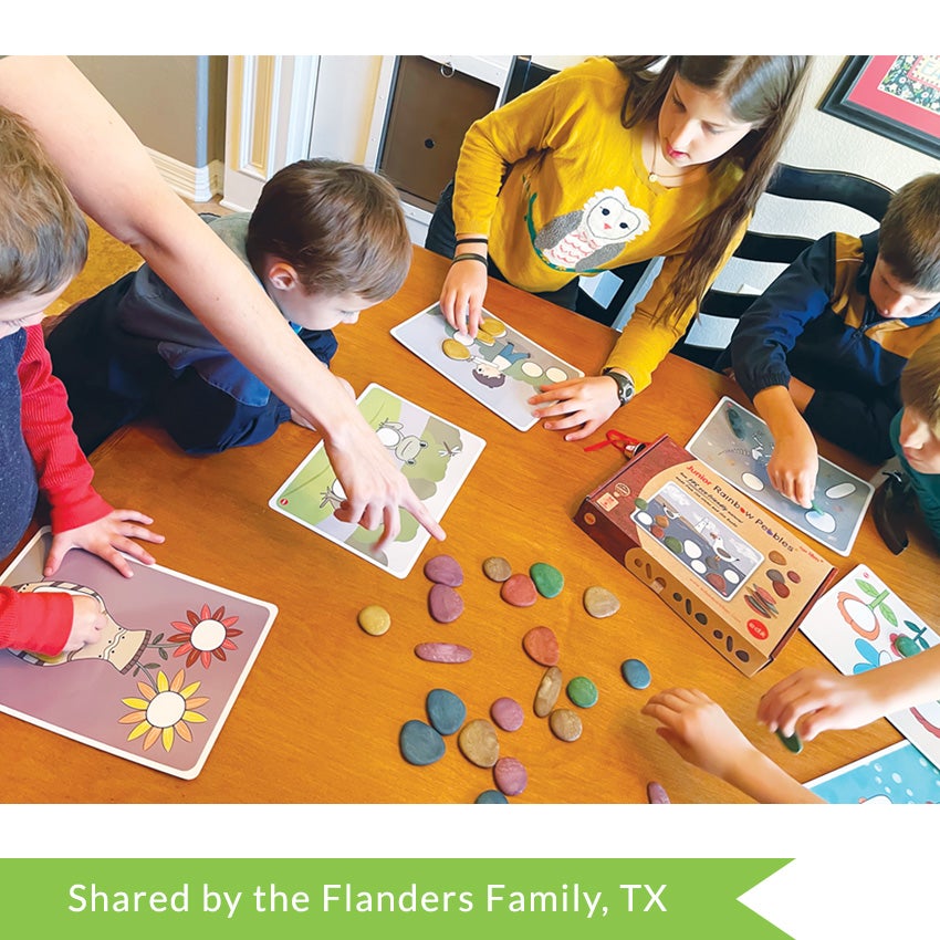 A customer photo of a family at their dining table, all playing with the Junior Rainbow Pebbles, Earth Tones. There is an adult hand pointing to the pebbles in the middle of the table and 6 children sitting around the table, each with an activity card in front of them and placing a pebble on top.