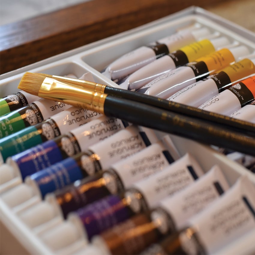 A close up of the Two Stick Shadows Paint Kit paint tray with a paintbrush on top. The paints in the tray are a variety of greens, blues, browns, yellows, oranges, black, and white.