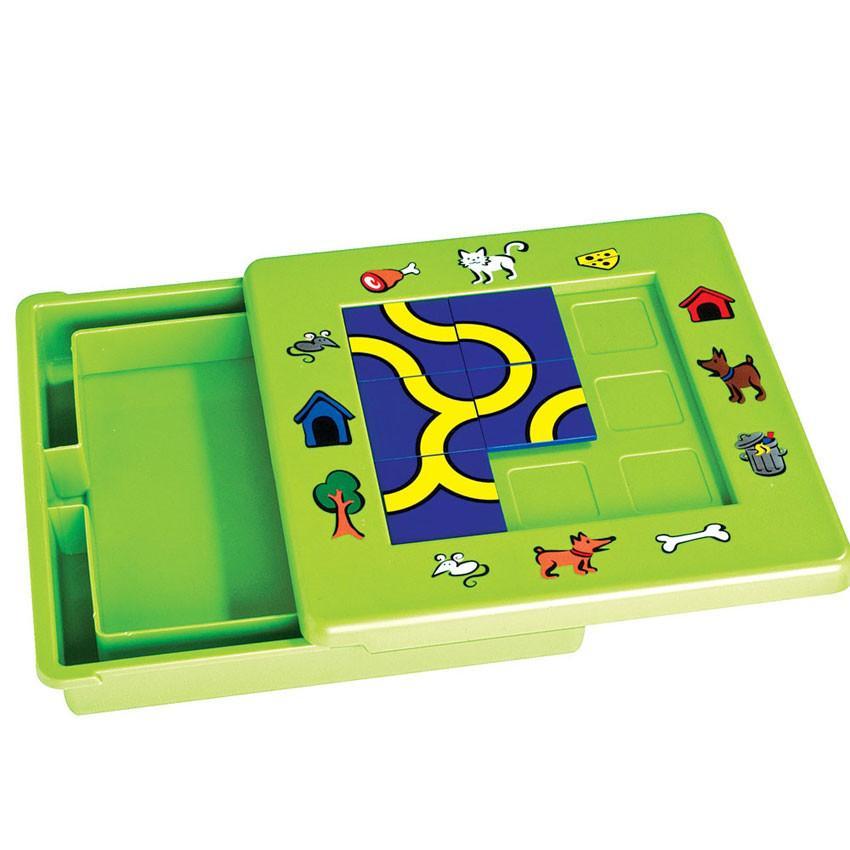 The Cat and Mouse game slid open to show the bottom of the board is storage for the playing pieces. The game board is bright green with illustrations around the board of pets, outside objects, pet snacks, and pet homes. There are blue and yellow square pathway pieces placed in the middle of the board, in a grid of 3 by 3.  There are only 5 of the 9 playing pieces placed on the board.