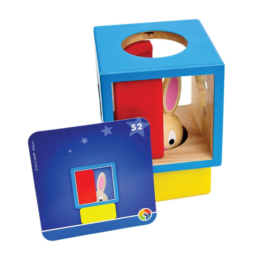The Bunny Boo game setup with a challenge card. The wood blocks are stacked with the yellow piece on bottom, then the hollow blue wood block with shapes cut out on the sides stacked on top. Tucked inside the blue block is a red block and the bunny tucked inside the red block, so you can only see half of his face. The blue challenge card is propped up against the blocks, showing the shapes stacked as they are.