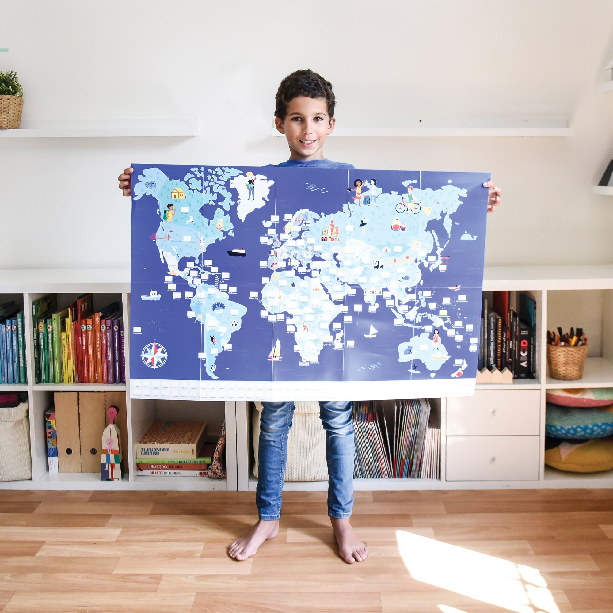 Dark-haired boy holding an outstretched world map with empty triangles for flag sticker placement. Child is wearing jeans with bare feet. In the background is a long, but short bookshelf with books and other items in front of a white wall.