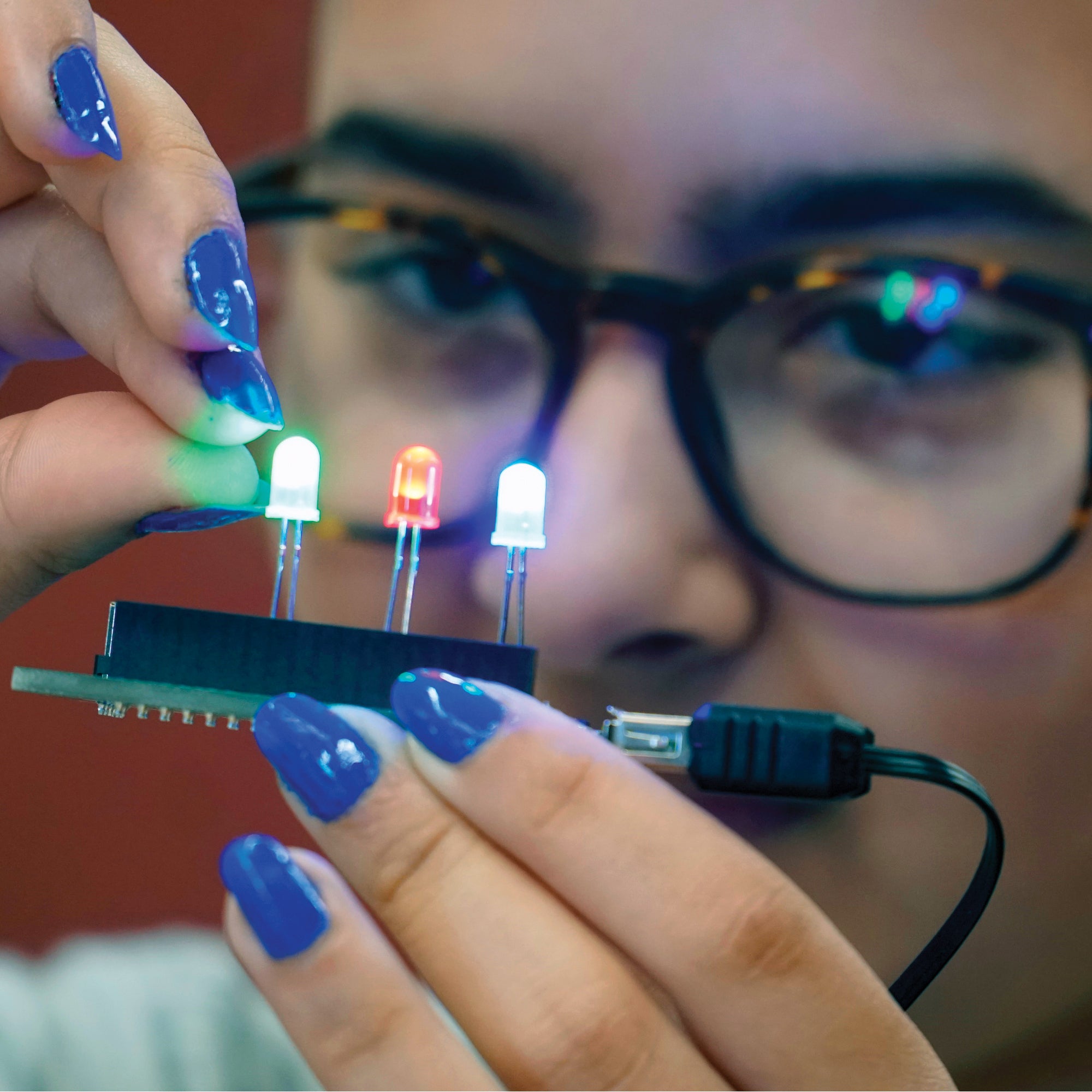 A close up of a teenage girl with glasses holding the Let’s Start Coding Maker (circuit) Board. She is holding the Maker Board in her left hand while her right hand is pinching toward the green L E D light attached. There is a green, red, and whitish-blue L E D light plugged in and lit up.