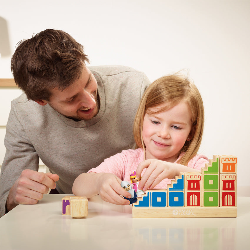 A father and daughter playing with the Camelot Junior game. The daughter is holding the knight and princess pieces with both hands and is walking them up the stairs on the side of a wood block piece. The other block pieces are stacked along the long base. The wood block pieces have castle walls painted on them in different colors. The father is leaning around from the back looking at his daughter playing with the pieces.