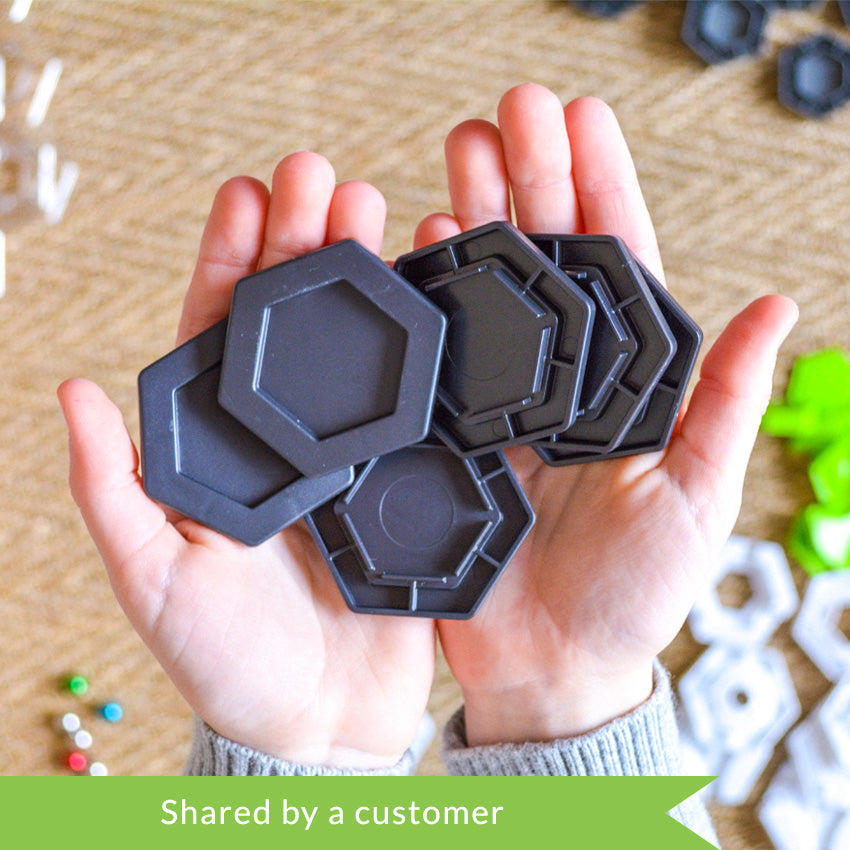 A customer photo from the top of a child’s hands holding 6 gray hexagon pieces from the GraviTrax set. Below the hands, and out of focus, are playing pieces from the set. The pieces shown are white hexagons, green hexagon attachment pieces, gray hexagons, colored marbles, and a clear hexagon plate. 