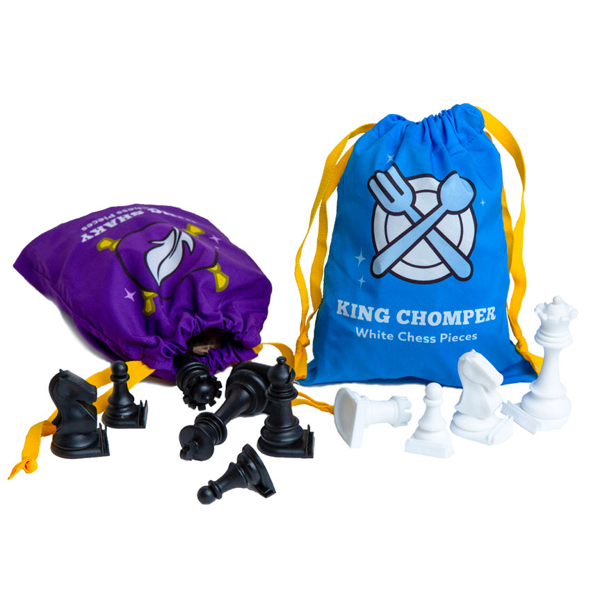 Story Time Chess storage bags on a white surface with pieces laid out in front. The left bag is purple with a white feather in the middle and laying down on the table. The pieces in and coming out of the bag are black. The standing blue bag on the right has a plate and crossed fork and spoon in the middle. There are white pieces in front. Both bags have yellow ribbon pull closures.