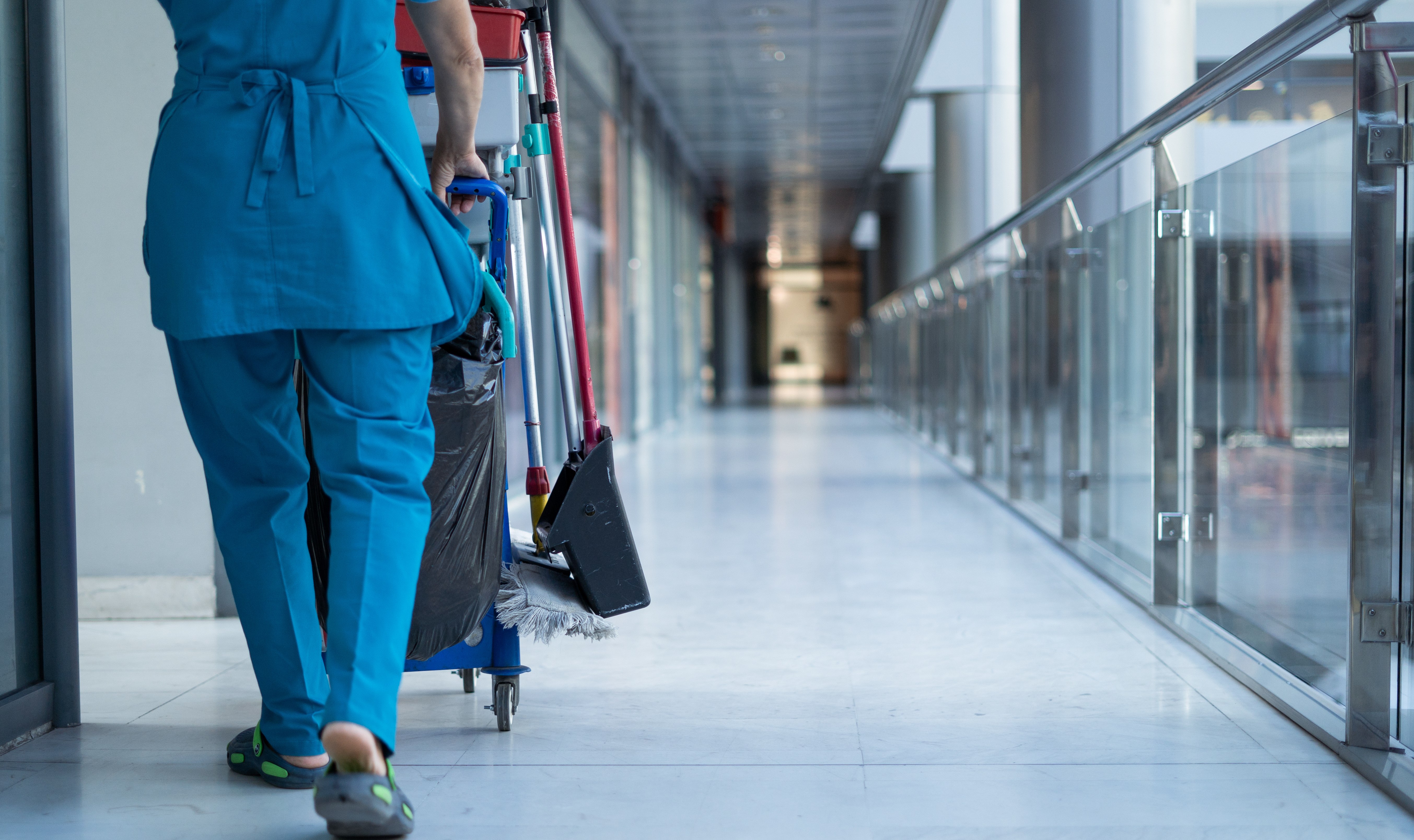 cleaning staff in blue uniform pushing cleaning cart down hallway in a sterile looking environment