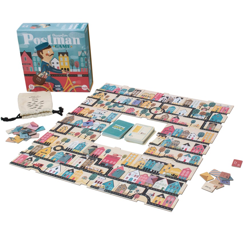 Postman Observation game laid out with the pieces. The board is a large square with a missing square in the middle. In the middle are 2 stacks of cards; one blue and one white. The board is covered in town buildings and houses in many colors. There are also roads and trees. On the sides of the board are some cut-out postal letter pieces. To the left is a storage bag and the game box. The box is blue with town buildings and a postman riding a bike.