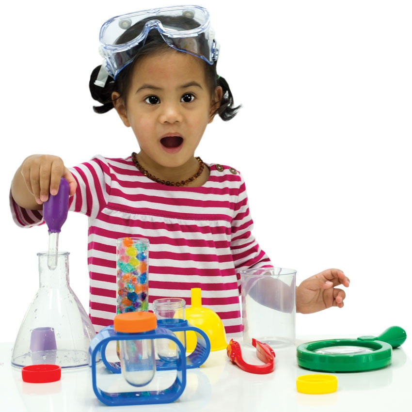 Dark-haired girl in a white and pink striped shirt. Her mouth is open in surprise. She is squeezing an eyedropper in her right hand into a beaker. You can see other contents of the Primary Science Lab Kit on the table in front of her including test tubes in stands, a yellow funnel, large red tweezers, a measuring cup, and a green magnifying glass.