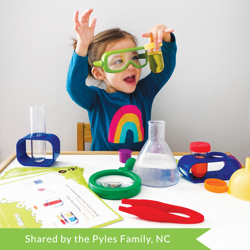 Customer photo of a brunette girl wearing green safety goggles and a dark teal shirt with a rainbow on it. She has her hands in the air, her left hand clutching a small test tube with yellow liquid inside. On the table in front of her are contents of the Primary Science Lab Kit including  activity cards, a tall test tube in a stand, a green magnifying glass, large red tweezers,  a beaker with clear liquid inside, a small test tube in a stand, and a yellow funnel.