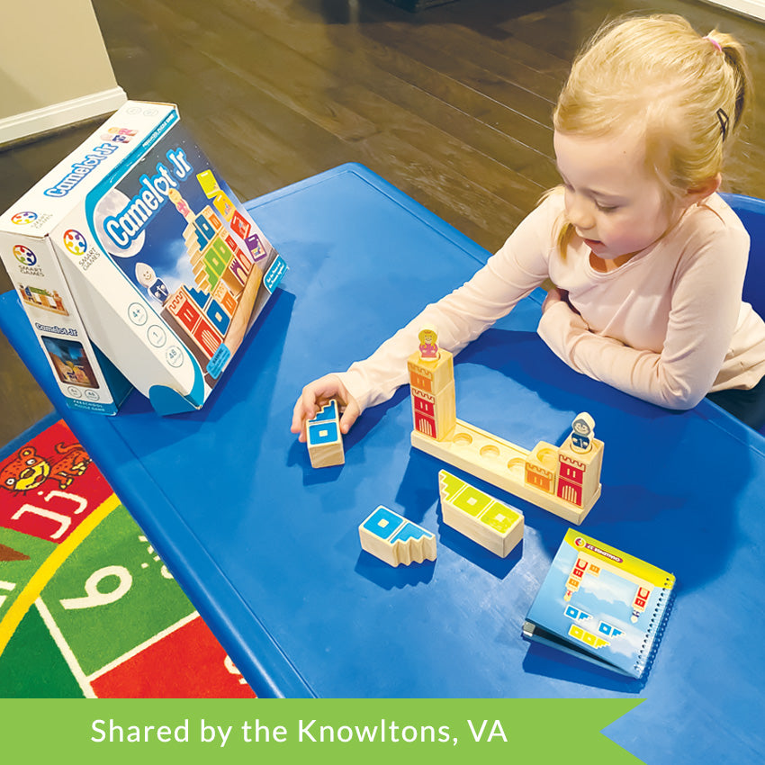 A customer photo of a young strawberry-blonde girl sitting at a blue table and playing with the Camelot Junior game. She is grabbing a wooden block to place it on the game base. The game base is a long, wooden, rectangle with blocks stacked on top. The wood block pieces have castle walls painted on them in different colors. There is a knight piece and a princess piece on top of wood block pieces. The instruction book is off to her left and the game box is off to her right.