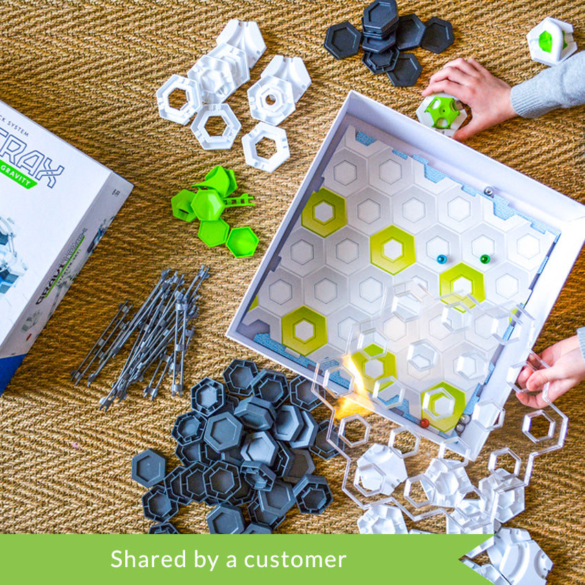 A customer photo from the top of a child’s hands playing with the GraviTrax set. The box is open with the cardboard base pieces and some marbles inside. Scattered around the box are piles of pieces. The pieces are dark gray hexagons, white hexagons, green hexagon attachment pieces, gray rails, gray hexagon pieces, and white hexagon pieces. The hands are holding a large clear hexagon piece to attach to the cardboard base.