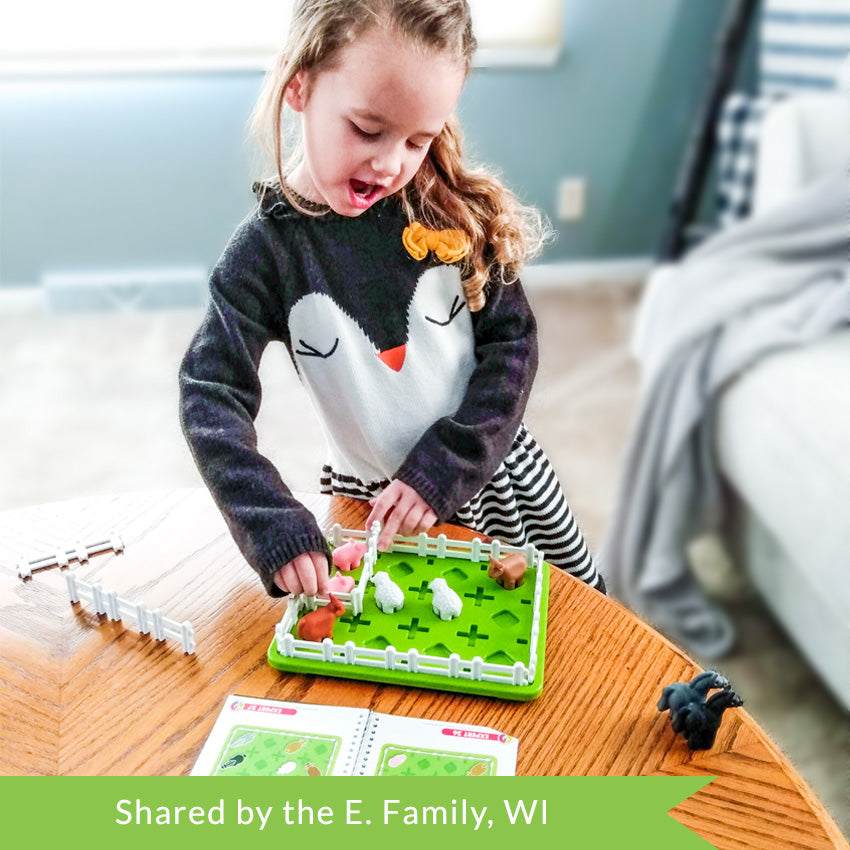 A customer photo of a young blonde girl in a penguin sweater playing with the Smart Farmer game. She has her hands on the game board, putting a white fence piece in place. The game board is green rectangle-shaped and has white fence pieces all around the edge. You can see 2 cows, 2 sheep, and 2 pigs inside the fence. Off to the side are 2 horses, fence pieces, and the instruction book open to show challenges.