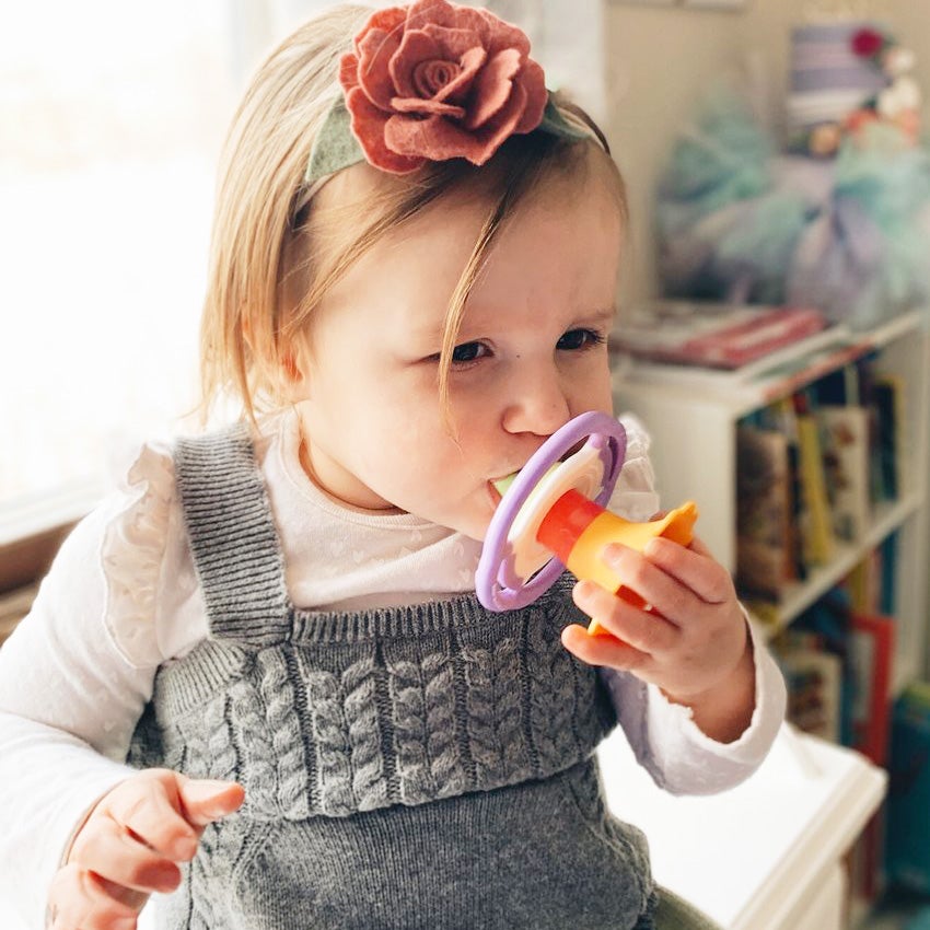 A blonde toddler wearing a flower headband is holding the Flower Whistle to her mouth with her left hand. The flower top of the whistle is a dark yellow with a pink center. The round middle portion of the whistle is white in the middle and lavender on the outside, where the handles are. The whistle portion at the bottom is bright green.