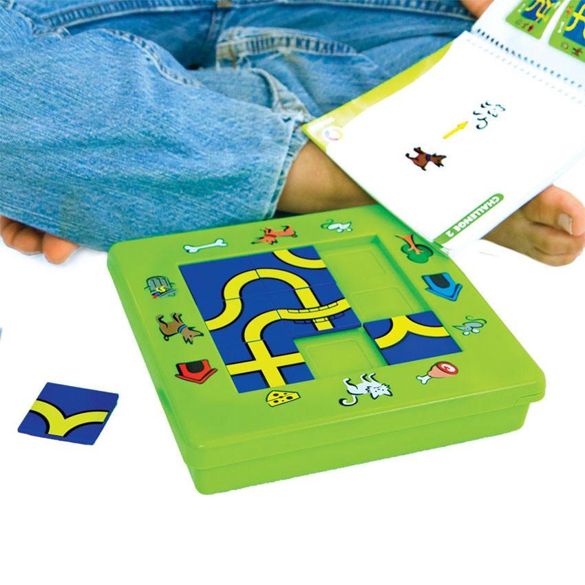 A close up shot of a boy playing The Cat and Mouse game on the floor. You see a boy sitting cross-legged with the instruction book on his foot. The game board in front of him is bright green with illustrations around the board of pets, outside objects, pet snacks, and pet homes. There are blue and yellow square pathway pieces placed in the middle of the board, in a grid of 3 by 3.  There are 6 of the 9 playing pieces placed on the board and 1 piece off to the side.