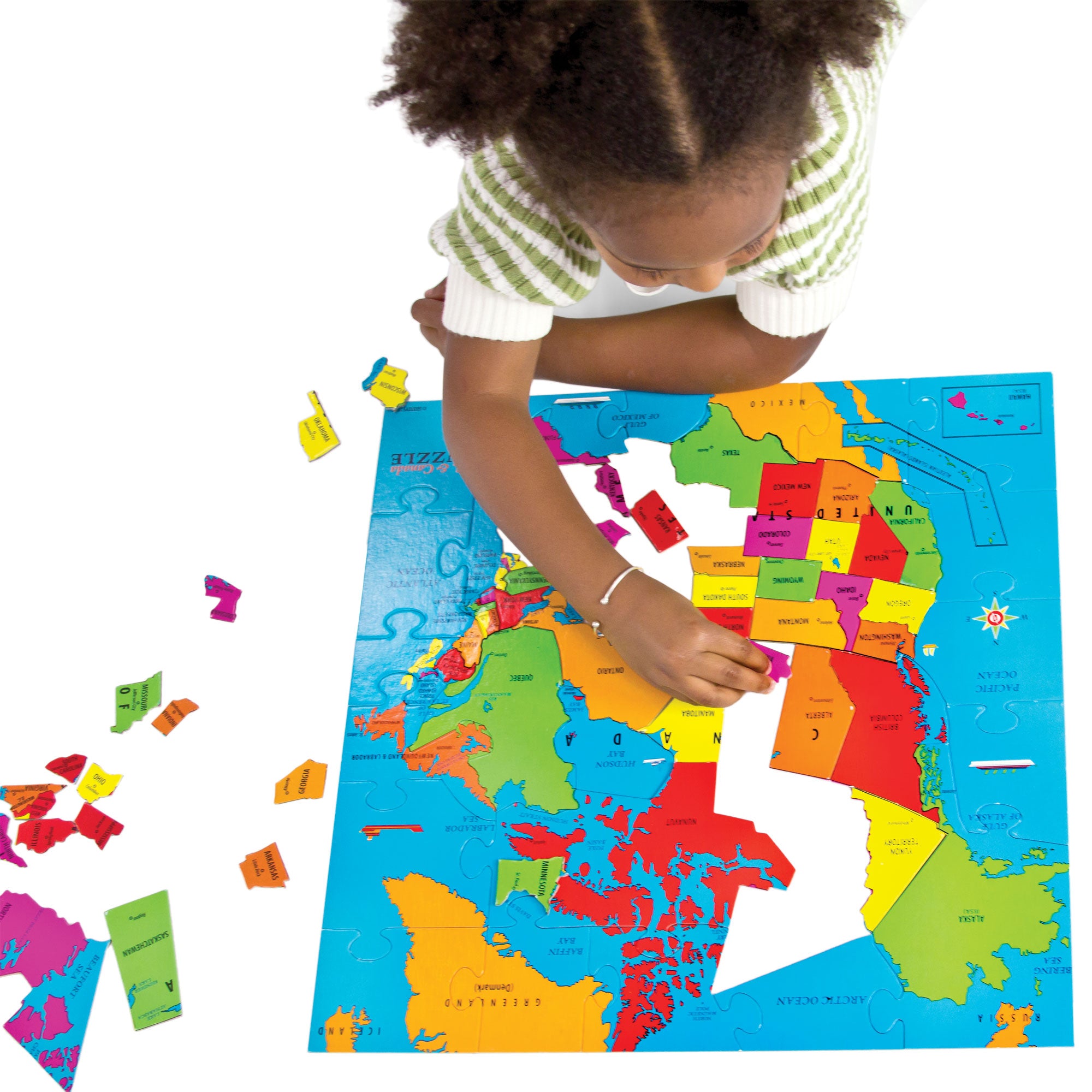 A young girl  with dark, fluffy, pig tails putting together the U S A and Canada Geo Puzzle. The puzzle is mostly put together with several pieces off to the left side. She is laying on the floor with her right hand stretched out putting in a piece. The continent shows the countries names and the names of their capitol. Each country is a different color. The colors are red, orange, yellow, green, and pink.