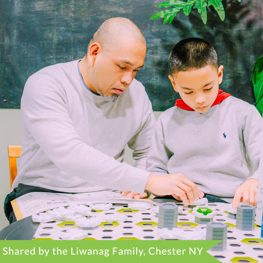 A customer photo of a father and son putting together the GraviTrax set. In front of them is a large white cardboard base to snap the pieces into. The base is white with green hexagons throughout. The pieces are mainly hexagon shapes in gray and white and are stacked in front of them with some white pieces in scattered piles on the base. The father is holding the instructions on his lap and placing a white piece on the board. The boy is also placing a white piece onto the board.