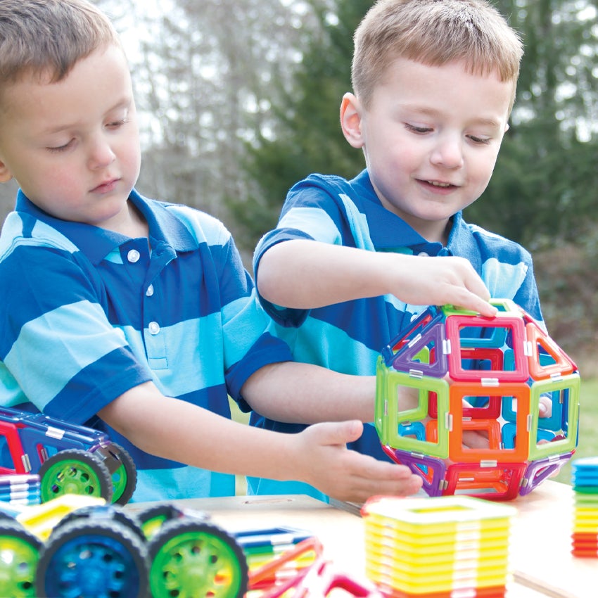 2 blonde boys in striped shirts are outside and are putting together a large shape, made up of the GeoSmart Educational Deluxe pieces on a picnic table. The pieces are geometrical shapes in squares and triangles and are a variety of different colors.  The boy on the left is holding a large rounded object in place on the sides, while the boy on the right is holding it in place on the top and the side. There are pieces in stacks and piles on the table, along with a car made from the pieces on the left.