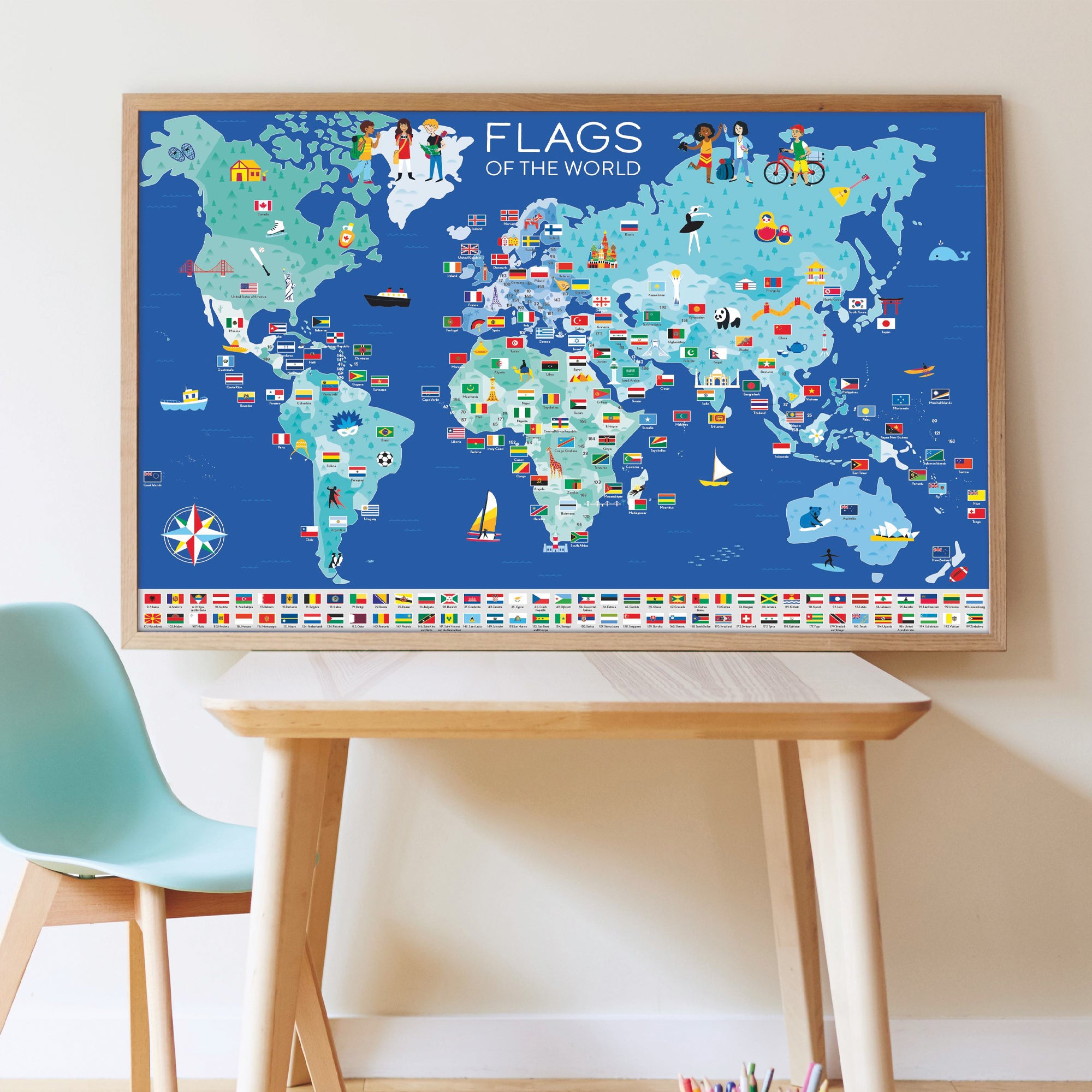 World map with flags framed and sitting on top of a light wood table with a turquoise chair off to the left and white walls behind.