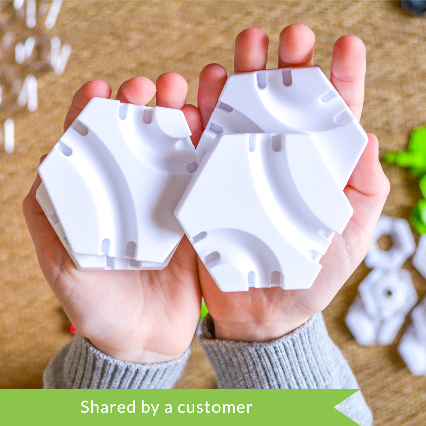 A customer photo from the top of a child’s hands holding 4 white hexagon pieces from the GraviTrax set. Below the hands, and out of focus, are playing pieces from the set. The pieces shown are white hexagons, green hexagon attachment pieces, colored marbles, and a clear hexagon plate. 