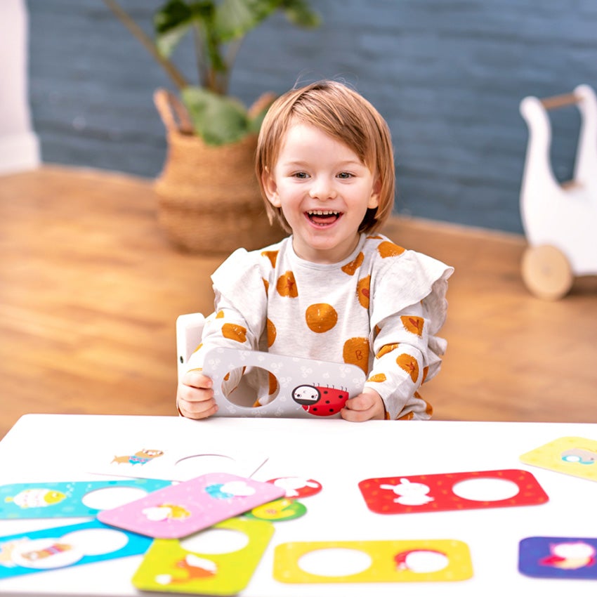 A customer photo of happy, smiling girl with short strawberry blonde hair. She is holding up the ladybug puzzle in front of the table she is sitting at. Strewn on the table are 9 more puzzles. The puzzles are rectangle shaped with rounded edges and a circle piece cut out on the right half.