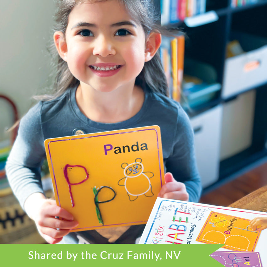 A customer photo of a brunette girl smiling and holding up a Wikki Stix Alphabet card. The orange card she is holding shows the letter P. It has an upper and lower case P with a  panda on it. On the table to her left is the box for the Wikki Stix Alphabet.