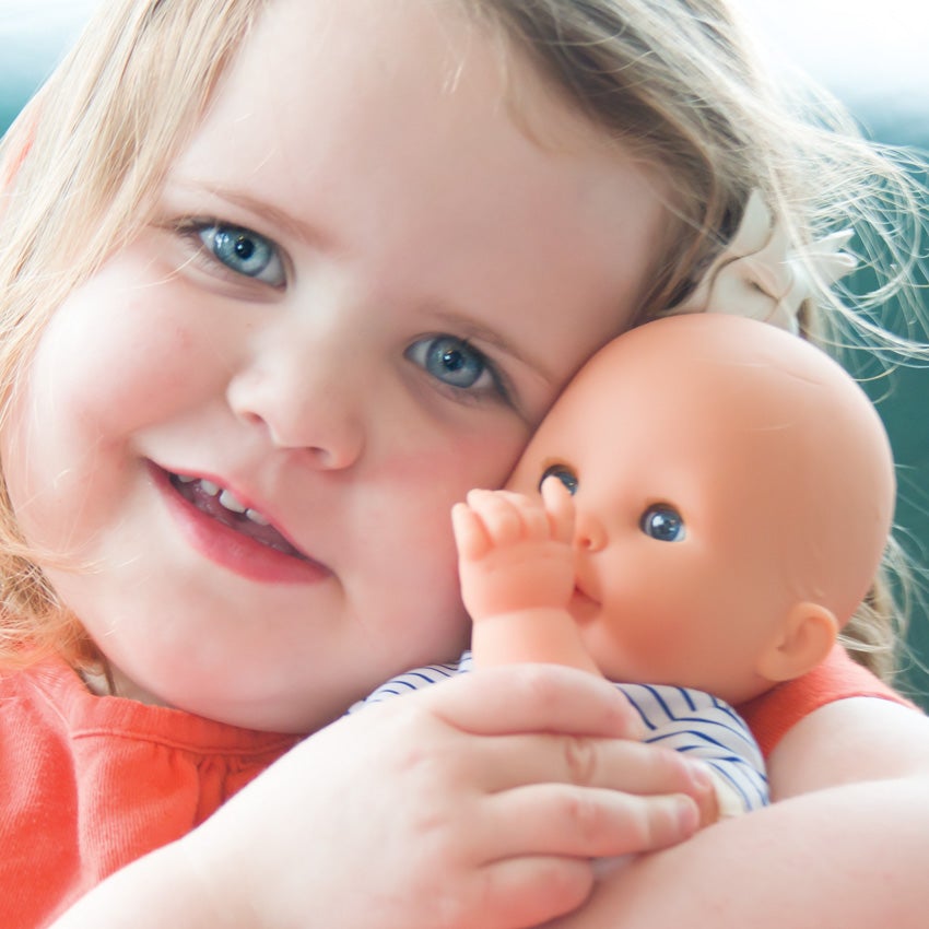 A close-up shot of a blonde, blue eyes girl swaddling Corolle’s Bebe Marin in her arms and smiling. The doll is light skinned with blue blinkable eyes. He is wearing a white shirt with blue stripes.