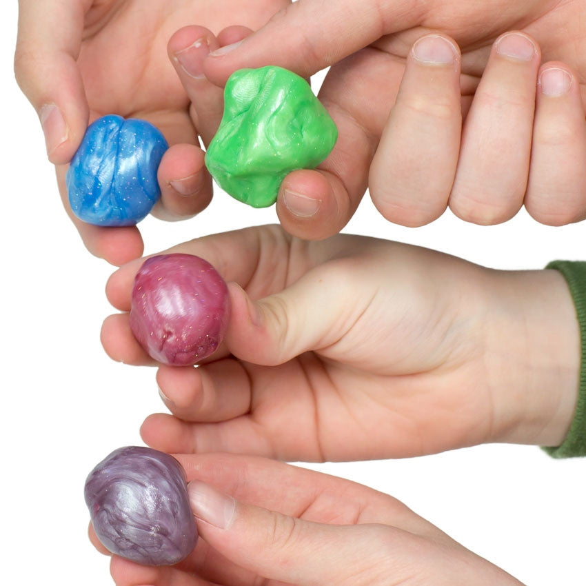A close-up of 4 children’s hands holding balls of Mixed by Me, Glow, Thinking Putty. The 2 hands at the top are holding glittery blue and glittery green putties. The next hand down is holding a glittery burgundy colored putty. The bottom hand is holing a lavender shimmer colored putty.