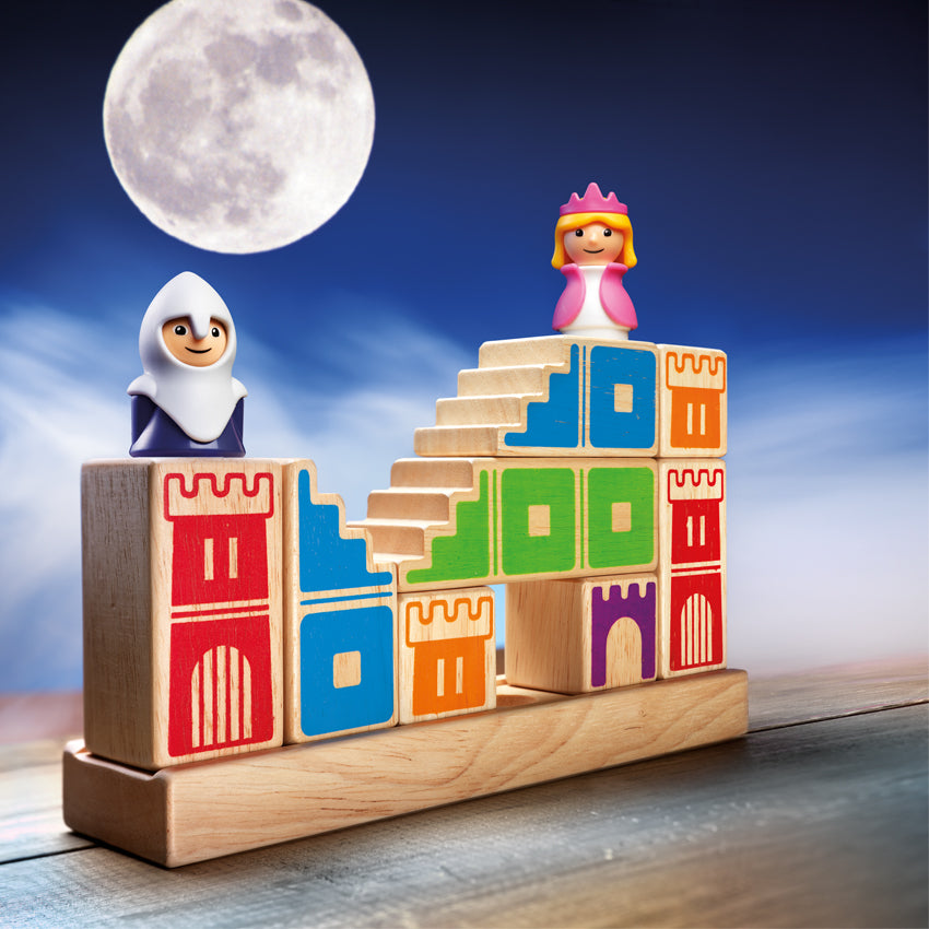 The Camelot Junior came setup with the knight and princess piece placed on top. The knight on the left is wearing a white and blue outfit. The blonde princess is wearing a pink and white outfit. The game base is a long, wooden, rectangle with blocks stacked on top. The wood block pieces have castle walls painted on them in different colors. In the background is a night sky with clouds and a large moon.
