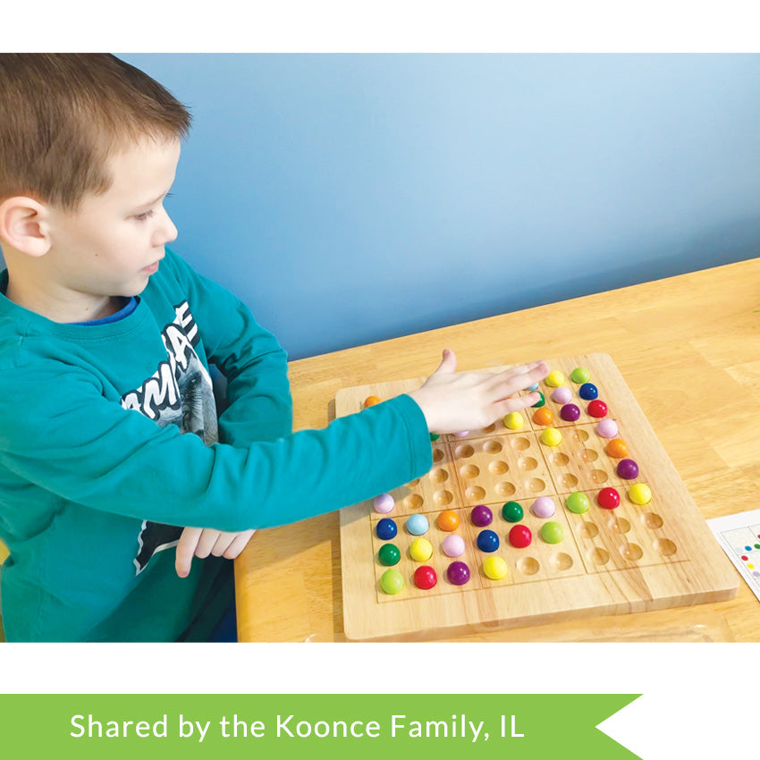 A customer photo of a brunette boy in the middle of playing ColorKu. He is looking down at the wooden board in front of him on the table and has his right hand touching one of the pieces on the board. The board in front of him has many pieces already in place. You can just make out a challenge card on the right side of the picture. The colored ball pieces are red, orange, yellow, bright green, dark green, light blue, dark blue, light purple, and dark purple.