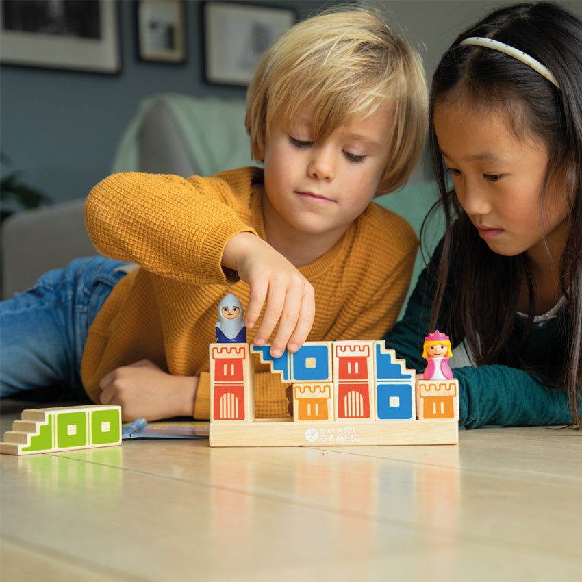 A young blonde boy and a dark-haired girl are playing with the Camelot Junior game while lying on the floor. The boy is stacking a wooden block on top of another and the girl is watching him put the piece in place. The base is a long, rectangle wood piece. The wood block pieces, stacked on top, have castle walls painted on them in different colors. There is a green painted wood piece and the instruction booklet off to the left in front of the boy.