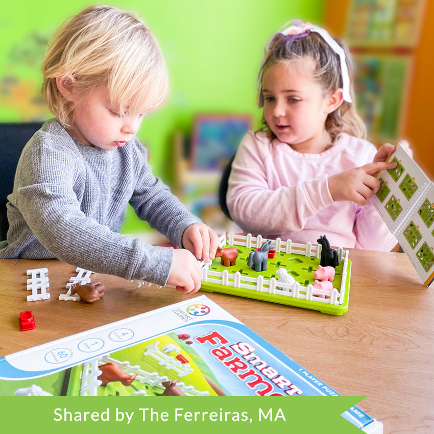 A customer photo of a young blonde boy and slightly older blonde girl playing with the Smart Farmer game. The boy is putting a fence piece onto the board with both hands. The girl, to the right, is holding up the instruction booklet and pointing out an instruction to the boy. The green, rectangle-shaped game board has white fence pieces all around the edge. You can see pigs, sheep, horses, and a cow inside the fence. Off to the side are more fence pieces, a cow, and a feeding dish.