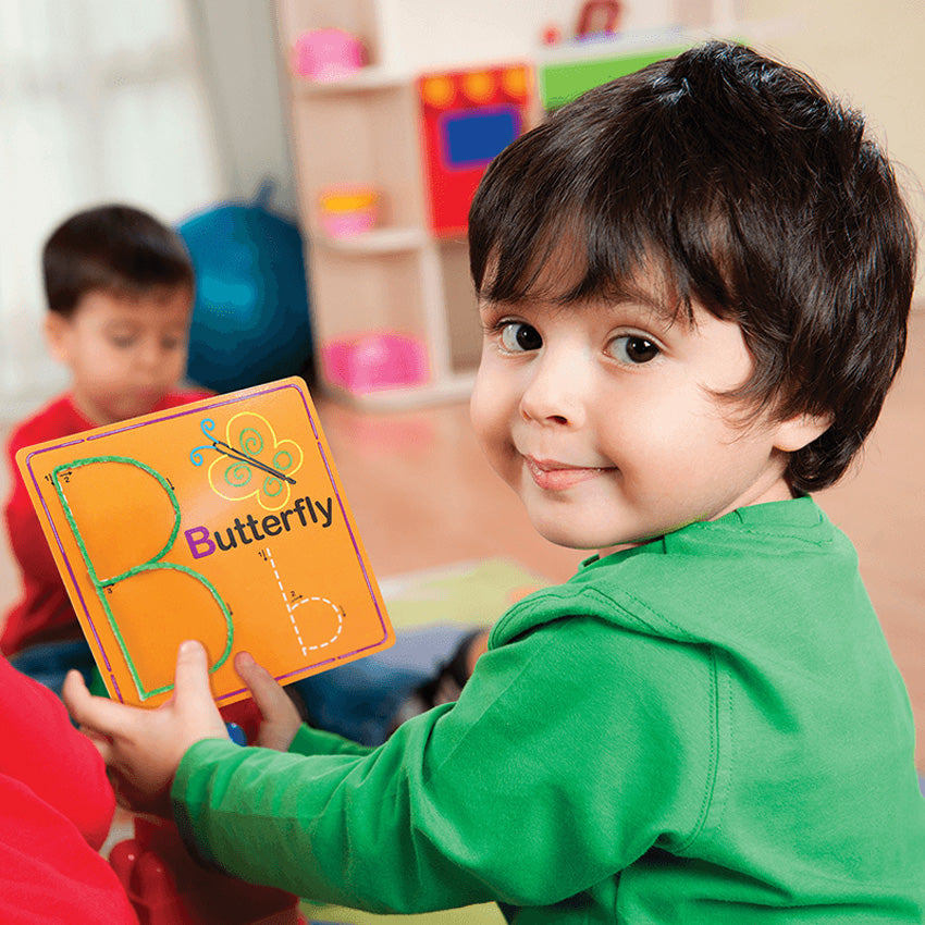 A young dark-haired boy in a green shirt is smiling and holding up a Wikki Stix Alphabet card. The orange card has a butterfly on it with an upper and lowercase b on it. The uppercase B is covered over with green wax sticks. You can see a boy playing in the background, out of focus.