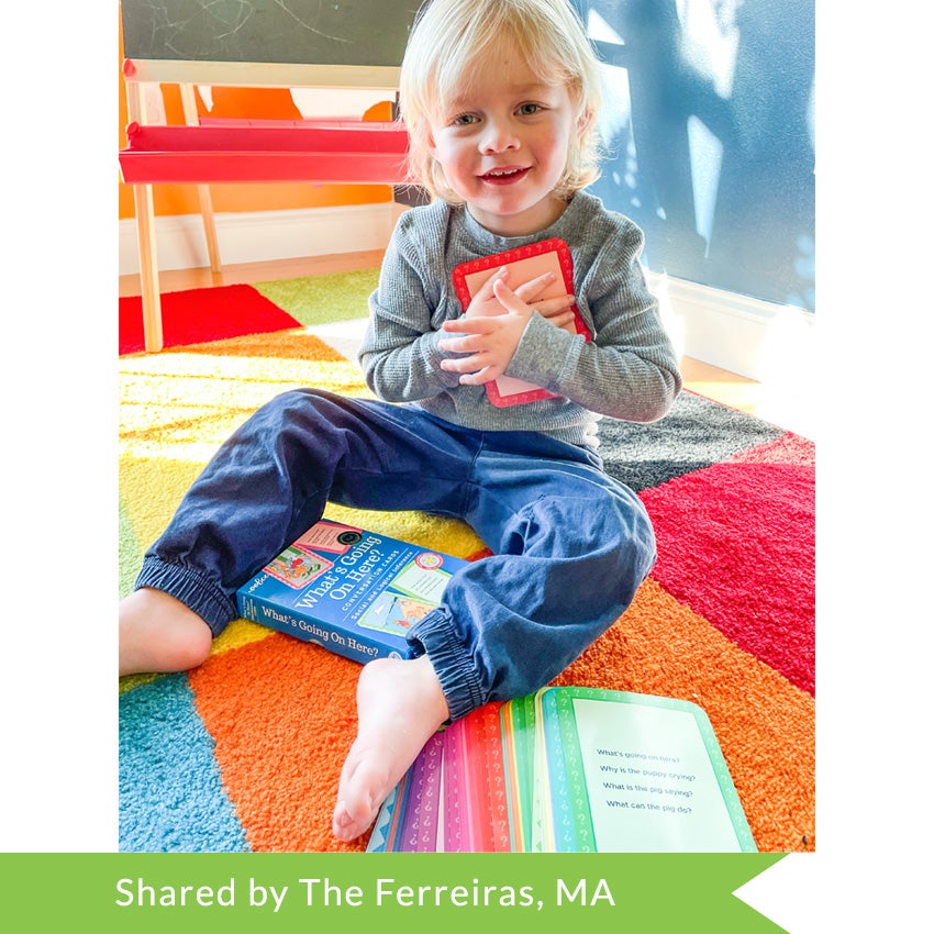 A customer photo of a blonde toddler sitting on the floor and smiling while holding a What’s Going on Here Conversation Card to his heart. The card is red and is large in his hands. Sitting between his legs is the card box. Toward the bottom of the picture, slightly under his foot, are more cards, of many colors, fanned out on the colorful square carpet.