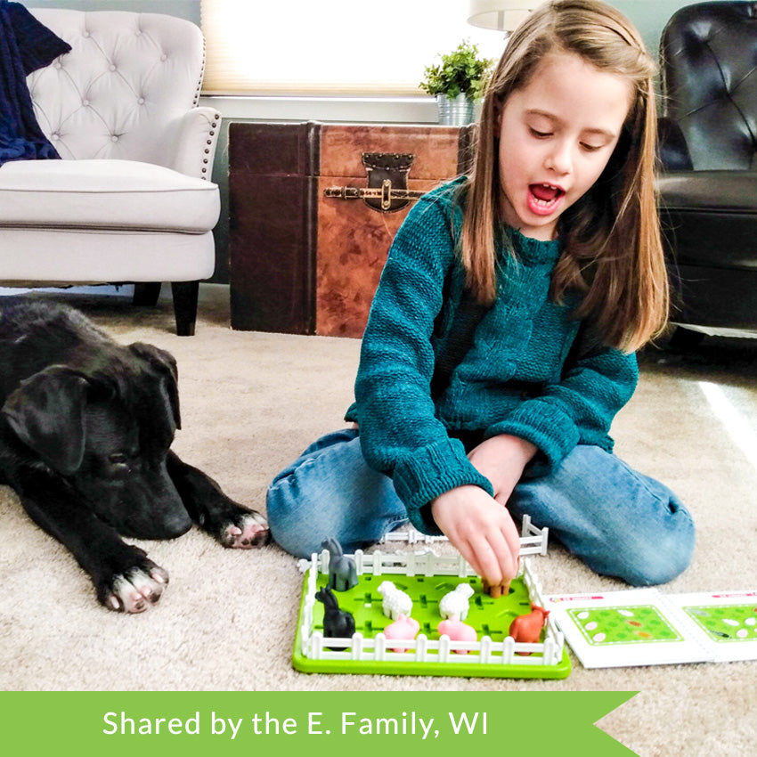 A customer photo of a young brunette girl playing with the Smart Farmer game next to her black dog on the floor. She is placing an animal piece on the board while talking. The green rectangle-shaped game board has white fence pieces all around the edge and a few through the middle. You can see 2 horses, 2 pigs, 2 sheep, and 2 cows inside the fence on the board. The instruction booklet is off to her left, open to show challenges.