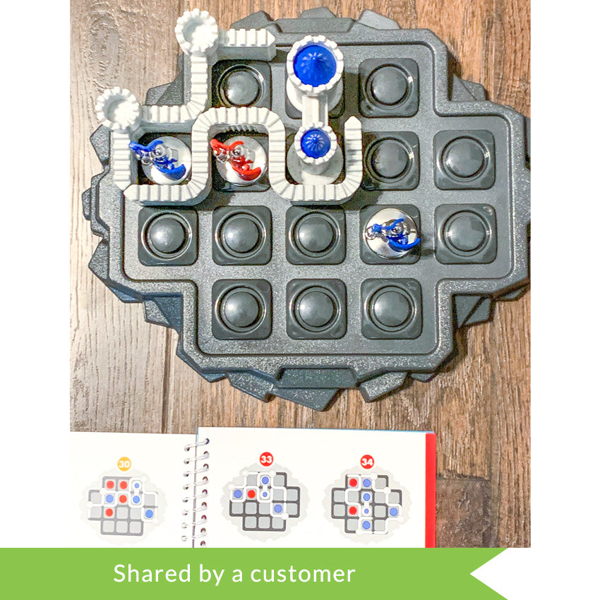 A customer photo of the Walls & Warriors game in play on a wood floor. The game base looks like a gray rock island with circles cut into squares on the top, allowing game pieces to be put in place. There are castle wall pieces and silver knight pieces with red and blue flags and shields placed on top and a game booklet open to show challenges off to the bottom-left.
