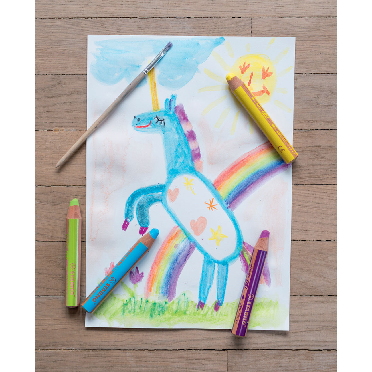 Wooden floor background with a watercolor of a unicorn rearing in front of a rainbow with a happy sun in the sky. 4 Stabilo pencils in yellow, purple, blue, and green placed on top of the picture along with a paintbrush.