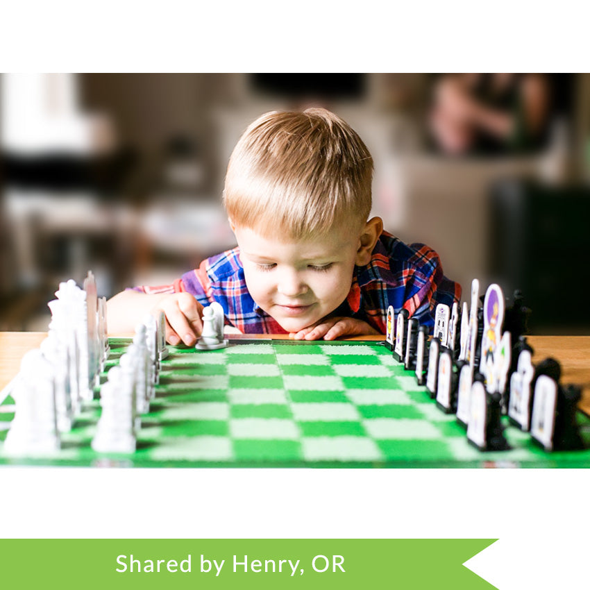 A customer photo of a young blonde boy resting his head on his hand that is on the table in front of him, planning his move. In front of him is the Story Time Chess game board. The game board squares are green grass, alternating darker green and lighter green. The pieces are standard chess shape, but has a character cut-out attached to each piece.