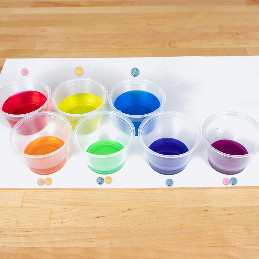 On top of a wood table are Color Fizzers laid out on a white paper with an accompanying glass with colored water. Above or below each glass shows which tablet, or tablets, make that color. The colors in the glasses are red, yellow, blue, orange, green, dark violet, and purple.