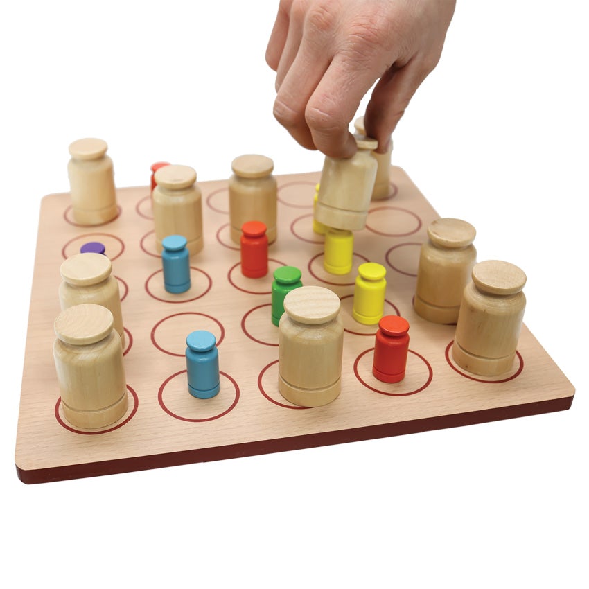 The Kloak game in play with a hand coming from the top and reaching down to grab a cover piece that is over a yellow piece. The square board is wooden with rounded corners and a grid of 5 by 5 circles on top. The pieces on top are colored wooden pieces that are rectangular with a tip shaped like a knob. The cover pieces are the same shape, but larger, and are wood colored.