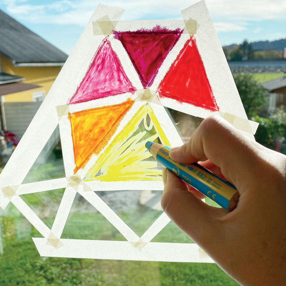 A hand holding a Stabilo pencil coloring on a window.  The window has masking tape placed to divide sections into triangles that are partially colored in with a variety of colors. You can see a neighborhood in the background, slightly blurry.