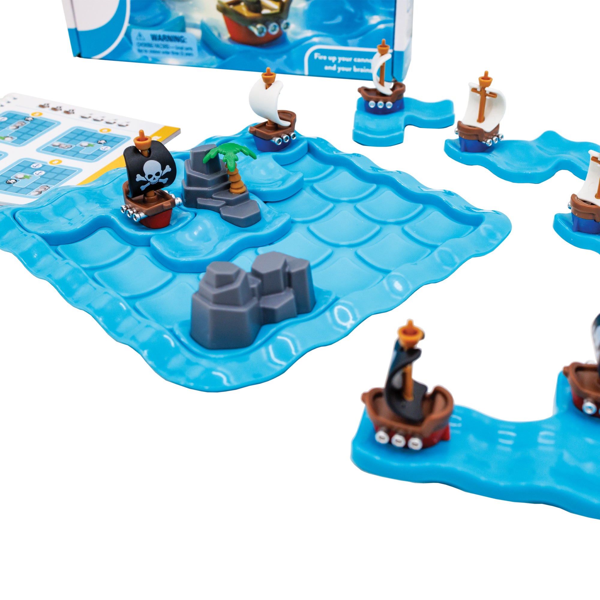 The Pirates Crossfire game. The gameboard is blue and there are 3 pieces set in place on the board; one ship with a white skull on a black sail, a rock piece, and another rock piece with a palm tree. You can see 4 ship pieces off to the right and the instruction book off to the left. You can see the bottom of the game box in the background, out of focus.