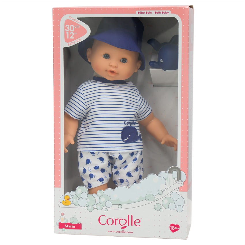 Corolle’s Bebe Marin standing in the box. Through the box window, you can see the light skinned baby doll has blue blinkable eyes. He is wearing shorts, a shirt, and a blue bucket hat. His shirt is white with blue stripes. His shorts are white with a blue whale pattern. In the top-right of the box is a rubber whale toy. At the bottom of the box is an illustration printed on the cardboard of a bathtub with streaming water and bubbles all around with a yellow rubber duck on the back.