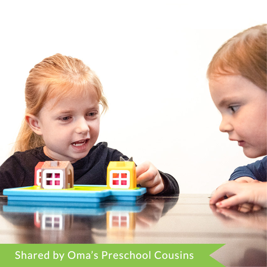A customer photo of 2 young, red-haired children playing with the Three Little Piggies game. They are sitting at a kitchen table. The girl on the left is placing the wolf piece onto the game board in front of her. The game board is light blue with house and pig pieces on the top. The boy on the right is looking intently at the game board as the girl plays.
