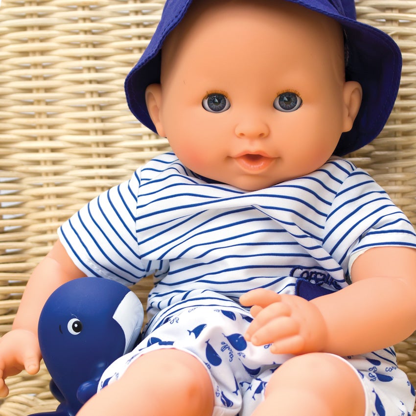 Corolle’s Bebe Marin. The light skinned baby doll has blue blinkable eyes and is sitting next to a rubber whale toy. He is wearing shorts, a shirt, and a blue bucket hat. His shirt is white with blue stripes. His shorts are white with a blue whale pattern. He is sitting on a tan wicker chair.